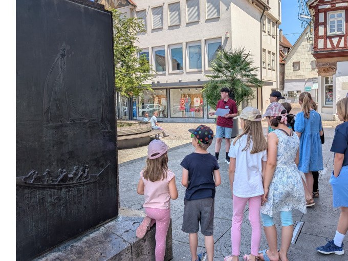 Kindergruppe hört einem jungen Mann in einer Stadt zu. Im Hintergrund sind Gebäude und ein Denkmal sichtbar., © Eigenbetrieb Tourismus und Citymanagement Schorndorf