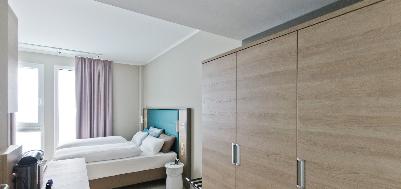 Ein modernes Hotelzimmer mit einem Doppelbett, einem großen Kleiderschrank und zwei Fenstern mit Vorhängen. Ein Tisch mit Wasserkocher ist sichtbar., © Arthotel Ana / elaya
