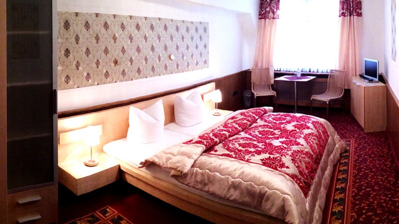 Gemütliches Hotelzimmer mit Doppelbett, rotem Teppich, Vorhängen, kleinem Tisch, zwei Stühlen und Fernseher.
