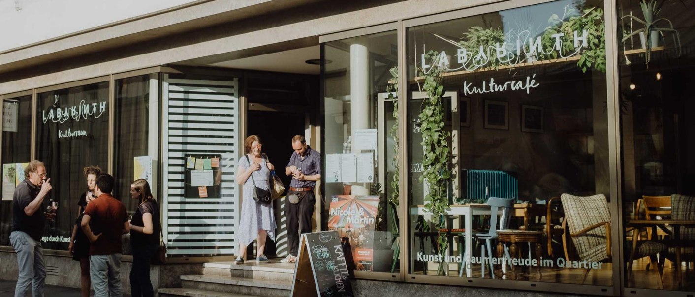 Menschen unterhalten sich vor dem LABYRINTH Kulturzentrum & -café. Die Fenster sind mit Pflanzen dekoriert, und ein Schild steht vor dem Eingang., © Christoph Steinweg