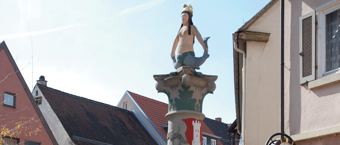 Eine Meerjungfrau mit Krone sitzt auf einem Brunnen, umgeben von Häusern mit roten Dächern., © Stuttgart-Marketing GmbH