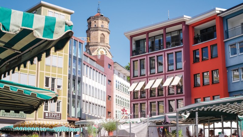 Bunte Gebäude und Marktstände auf einem Marktplatz, mit einem Kirchturm im Hintergrund unter blauem Himmel., © SMG, trickytine