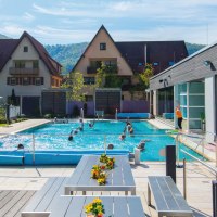 Thermalbad mit Pool, Liegestühlen und Sonnenschirmen. Menschen schwimmen im Wasser, im Hintergrund sind Häuser und grüne Hügel zu sehen., © SMG Achim Mende