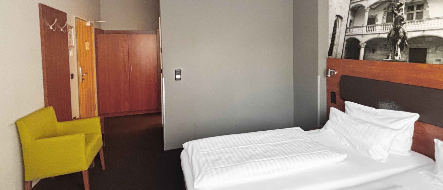 Ein Hotelzimmer mit zwei Einzelbetten, einem gelben Sessel und einem gro&szlig;en Schrank. An der Wand h&auml;ngt ein Schwarz-Wei&szlig;-Foto eines historischen Geb&auml;udes., &copy; Novum Hospitality