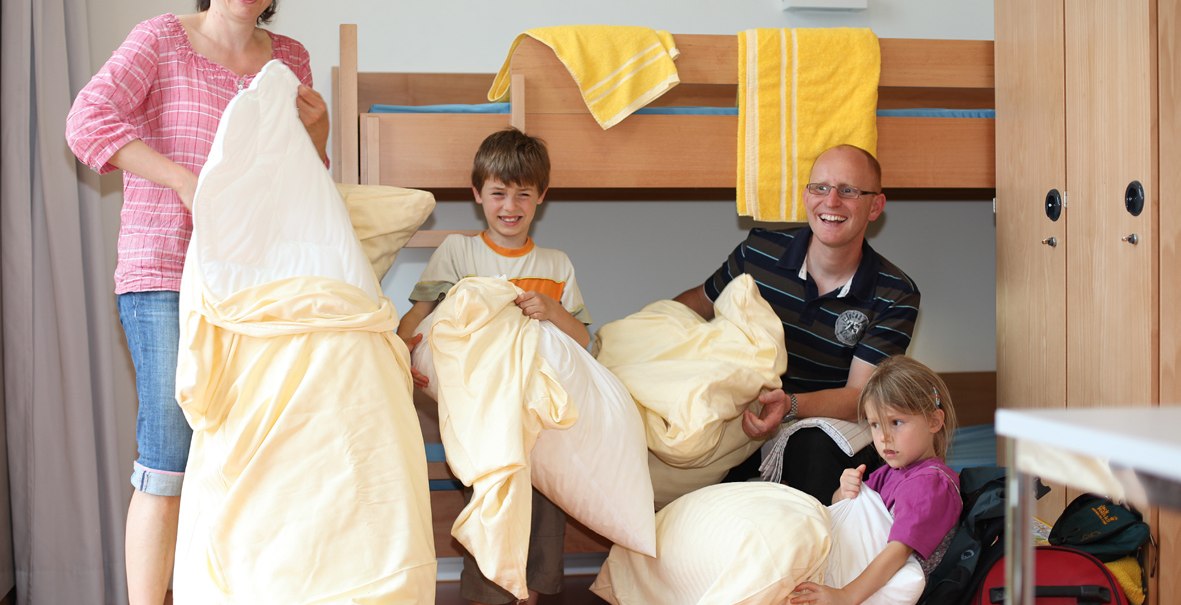 Eine Familie in einem Zimmer mit Etagenbett. Sie halten gelbe Bettwäsche und lächeln. Handtücher hängen am Bett. Ein Kind sitzt auf einem Koffer., © DJH Landesverband Baden-Württemberg e.V.