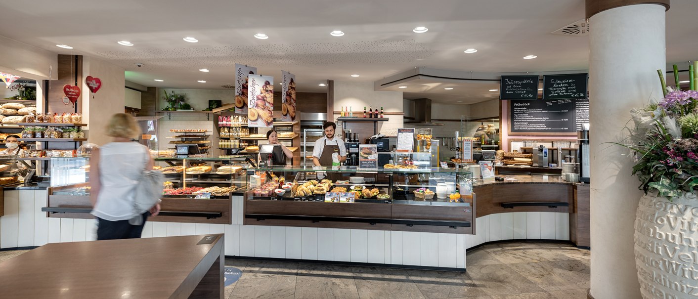 Eine Bäckerei mit Theke voller Backwaren. Zwei Mitarbeiter bedienen Kunden. Die Einrichtung ist modern und einladend., © Trölsch GmbH