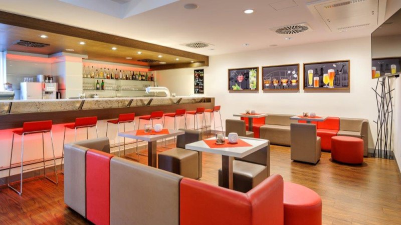 Moderne Bar mit roten und grauen Möbeln, beleuchtetem Tresen und Wandbildern von Getränken. Elegantes Ambiente mit Holzboden., © Ibis Stuttgart Airport Messe Moderne Bar mit roten und grauen Möbeln, beleuchtetem Tresen und Wandbildern von Getränken. Elegantes Ambiente mit Holzboden., © Ibis Stuttgart Airport Messe