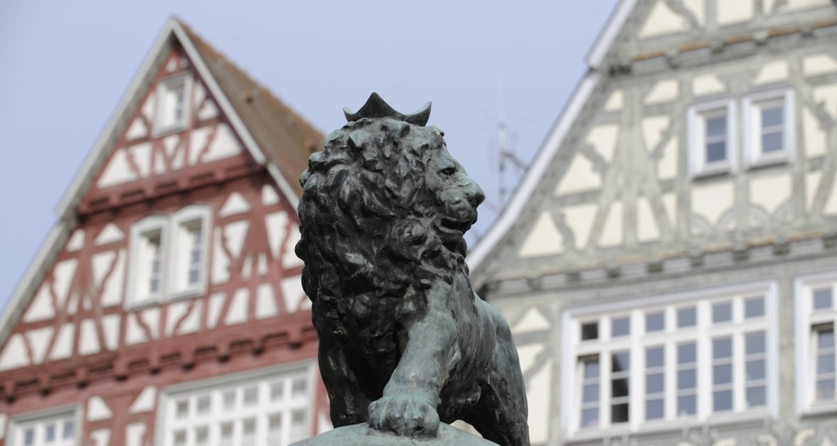 Eine Bronzestatue eines Löwen mit Krone steht vor zwei Fachwerkhäusern. Die Häuser haben weiße und rote sowie weiße und graue Fassaden., © Land der 1000 Hügel - Kraichgau-Stromberg Eine Bronzestatue eines Löwen mit Krone steht vor zwei Fachwerkhäusern. Die Häuser haben weiße und rote sowie weiße und graue Fassaden., © Land der 1000 Hügel - Kraichgau-Stromberg