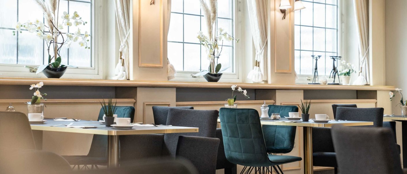 Elegantes Frühstückszimmer mit gedeckten Tischen, modernen Stühlen und großen Fenstern. Dezente Dekoration mit Pflanzen und stilvollen Lampen., © Flairhotel Vier Jahreszeiten