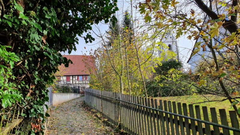 Ein gepflasterter Weg führt durch eine grüne Umgebung zu einem Fachwerkhaus und einem Kirchturm. Herbstliche Blätter und ein Holzzaun säumen den Weg., © Kulturamt Plochingen Ein gepflasterter Weg führt durch eine grüne Umgebung zu einem Fachwerkhaus und einem Kirchturm. Herbstliche Blätter und ein Holzzaun säumen den Weg., © Kulturamt Plochingen