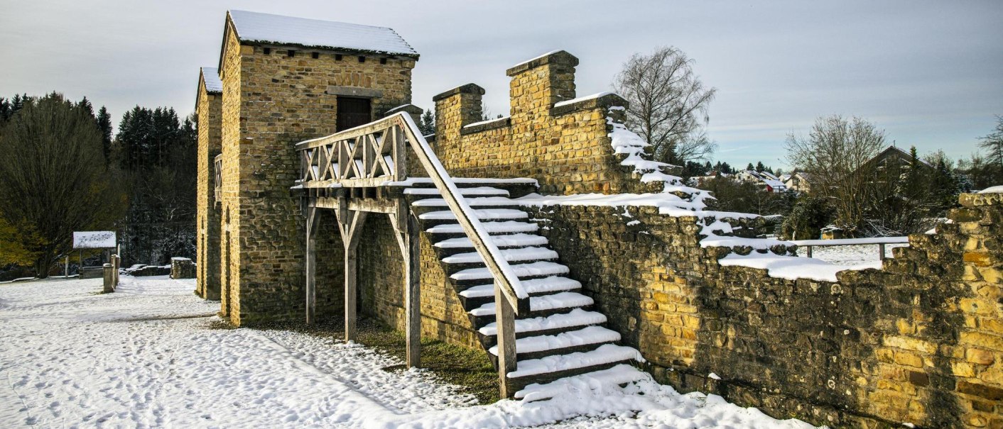 Das Römische Ostkastell ist mit Schnee bedeckt. Eine Holztreppe führt zu den alten Steinmauern. Im Hintergrund sind Bäume und Häuser zu sehen., © SMG, Sarah Schmid