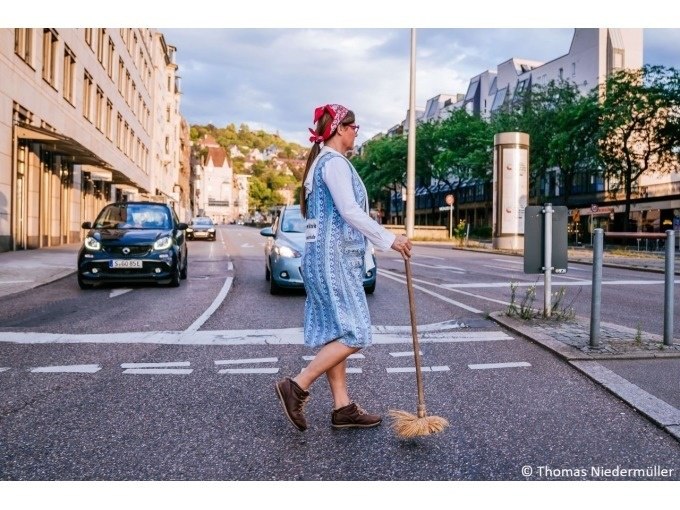Eine Frau in traditioneller Kleidung mit Kopftuch und Besen überquert eine Straße in einer Stadt. Autos sind im Hintergrund zu sehen., © Stuttgart Marketing GmbH Eine Frau in traditioneller Kleidung mit Kopftuch und Besen überquert eine Straße in einer Stadt. Autos sind im Hintergrund zu sehen., © Stuttgart Marketing GmbH