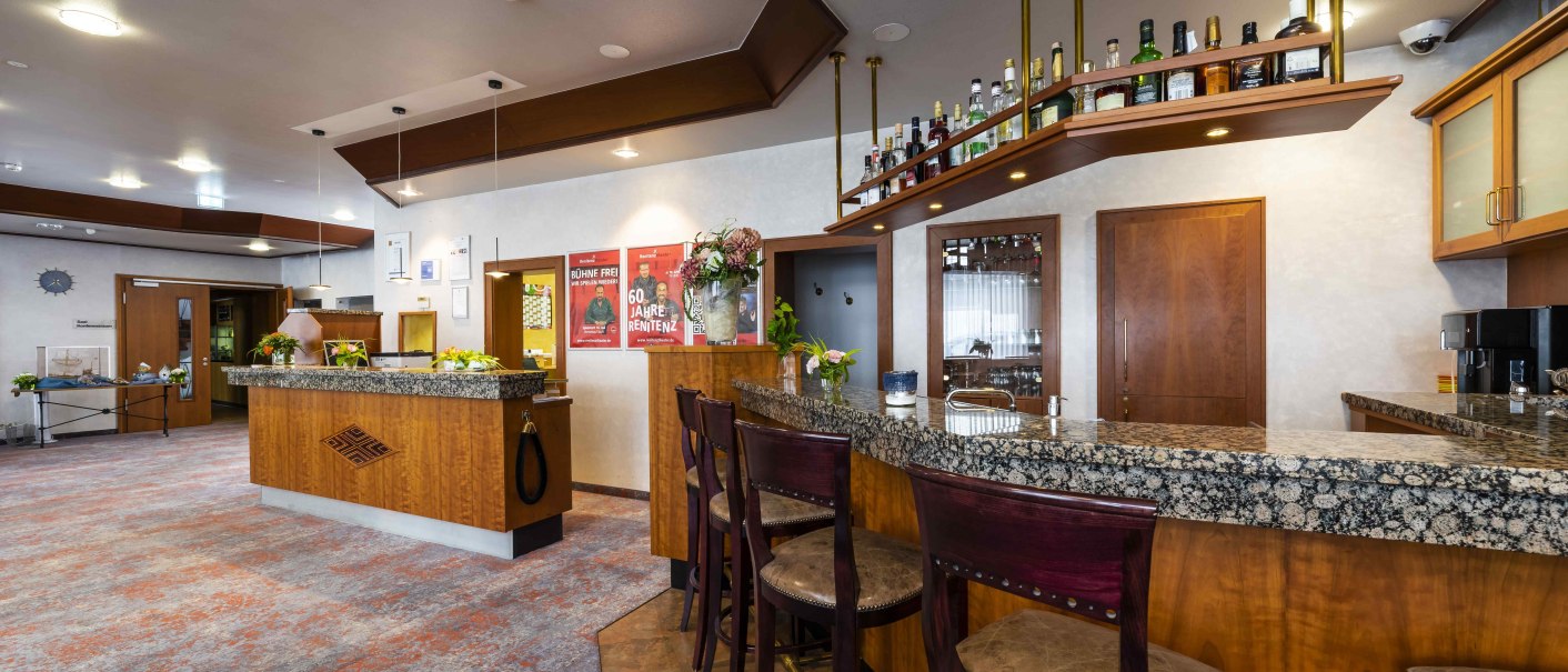 Hotelrezeption mit Bar, Holzmöbeln und gepolsterten Stühlen. Flaschen stehen auf einem Regal, Blumenarrangements dekorieren den Raum., © TOMAS