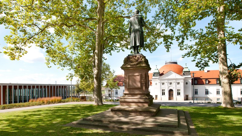 Statue auf der Schillerh&ouml;he in Marbach am Neckar, umgeben von B&auml;umen. Im Hintergrund ein historisches Geb&auml;ude mit rotem Dach., &copy; Corinna Jacobs