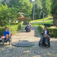 Drei Personen in Rollstühlen fahren auf einem gepflasterten Weg durch einen grünen Park mit Bäumen und Sträuchern., © NPSFW Drei Personen in Rollstühlen fahren auf einem gepflasterten Weg durch einen grünen Park mit Bäumen und Sträuchern., © NPSFW