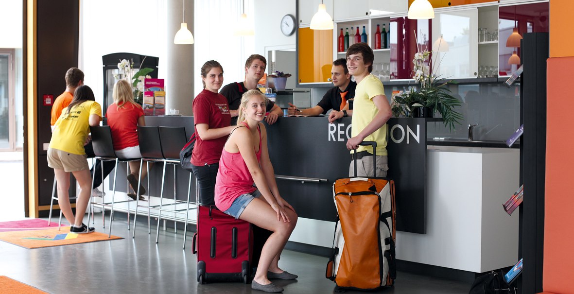 Junge Menschen mit Gepäck an einer modernen Hotelrezeption. Einige sitzen, andere stehen, während sie einchecken. Helle und freundliche Atmosphäre., © DJH Landesverband Baden-Württemberg e.V.