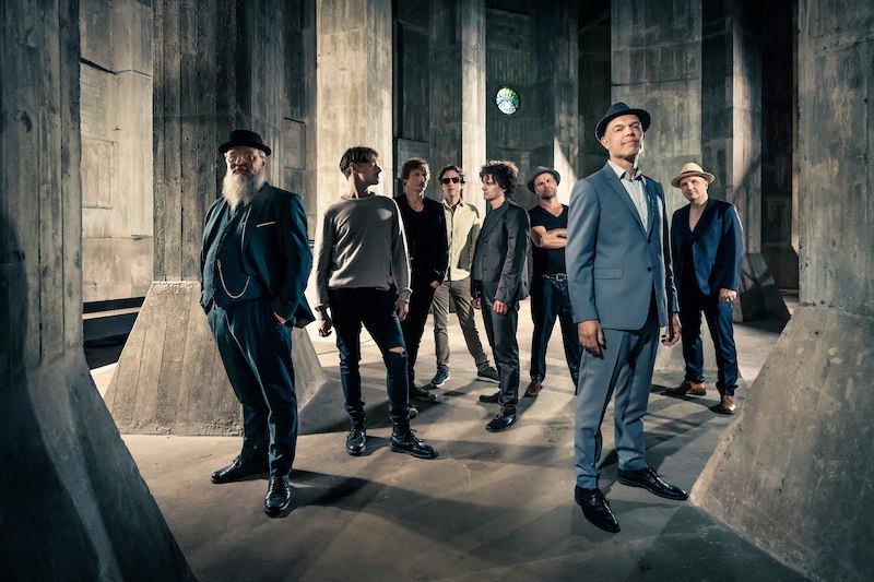 Acht Männer in eleganter Kleidung posieren in einem Raum mit Betonpfeilern. Einer trägt einen Hut, andere Sonnenbrillen. Die Atmosphäre ist industriell., © Scala Kultur Live gGmbH