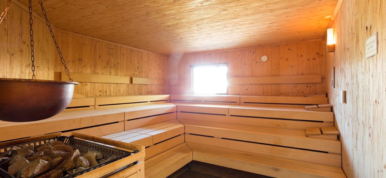 Holzverkleidete Sauna mit Bänken und einem Ofen mit Steinen, warmes Licht und kleines Fenster., © ARAMIS Hotel & Freizeit GmbH