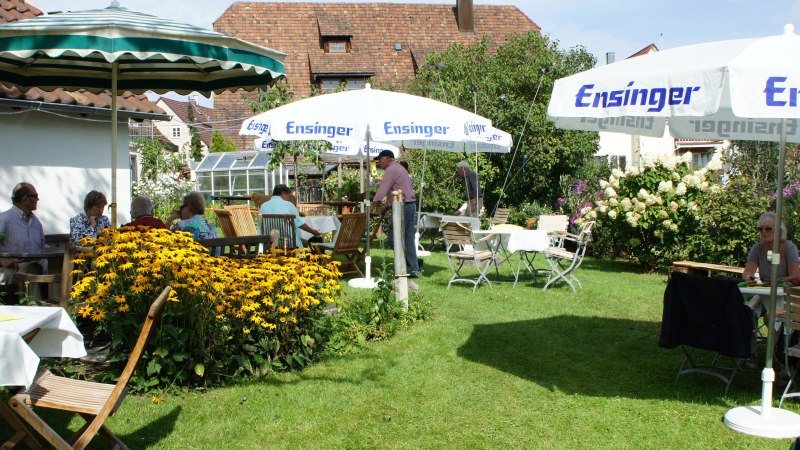 Ein sommerlicher Garten mit Menschen, die unter Sonnenschirmen an Tischen sitzen, umgeben von blühenden Blumen und grüner Vegetation., © Weinbau Knobloch-Wolfrum Ein sommerlicher Garten mit Menschen, die unter Sonnenschirmen an Tischen sitzen, umgeben von blühenden Blumen und grüner Vegetation., © Weinbau Knobloch-Wolfrum