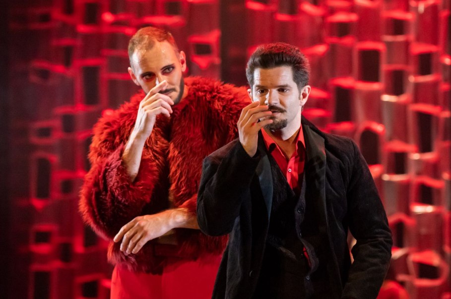 Zwei Männer in auffälliger Kleidung stehen auf einer Bühne. Der Hintergrund ist rot beleuchtet, was eine dramatische Atmosphäre schafft.