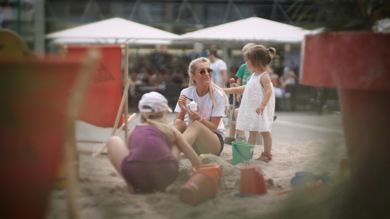 Eine Frau und zwei Kinder spielen im Sand an einem Stadtstrand. Die Frau isst Eis, w&auml;hrend die Kinder mit Eimern und Schaufeln spielen., &copy; Stadtmarketing G&ouml;ppingen
