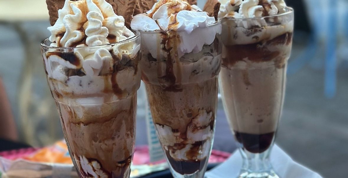 Drei hohe Gläser mit Eiskaffee, Sahne und Keksen auf einem Tablett. Im Hintergrund sind unscharfe Gartenmöbel zu sehen., © E. Dogan Drei hohe Gläser mit Eiskaffee, Sahne und Keksen auf einem Tablett. Im Hintergrund sind unscharfe Gartenmöbel zu sehen., © E. Dogan