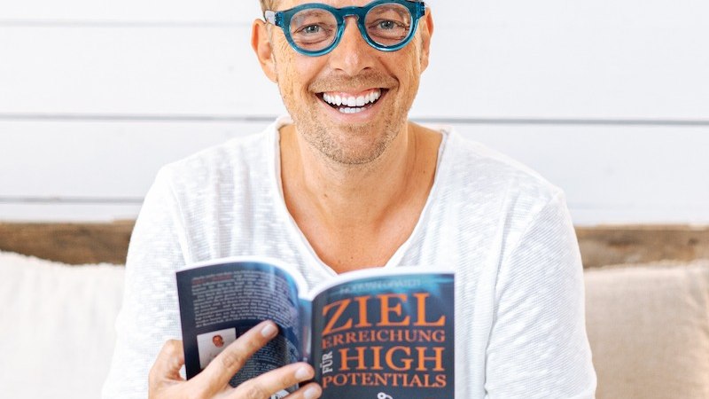 Ein Mann mit blauer Brille und wei&szlig;em Hemd h&auml;lt l&auml;chelnd ein Buch mit dem Titel 'Zielerreichung f&uuml;r High Potentials' in den H&auml;nden., &copy; Mercedes-Benz AG