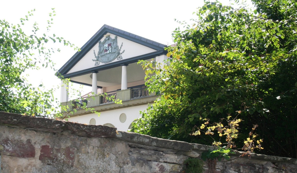 Ein Gebäude mit Säulen und einem Wappen auf der Fassade, umgeben von Bäumen und einer Steinmauer im Vordergrund., © Natur.Nah. Schönbuch & Heckengäu
