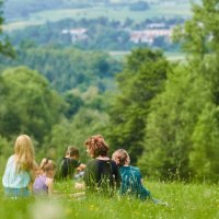 Gruppe von Menschen sitzt auf einer gr&uuml;nen Wiese, umgeben von B&auml;umen, mit Blick auf ein Dorf in der Ferne., &copy; Natur.Nah. Sch&ouml;nbuch & Heckeng&auml;u