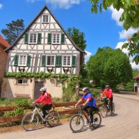 Drei Radfahrer in bunter Kleidung fahren an einem malerischen Fachwerkhaus vorbei. Der Himmel ist blau mit weißen Wolken., © Land der 1000 Hügel - Kraichgau-Stromberg Drei Radfahrer in bunter Kleidung fahren an einem malerischen Fachwerkhaus vorbei. Der Himmel ist blau mit weißen Wolken., © Land der 1000 Hügel - Kraichgau-Stromberg
