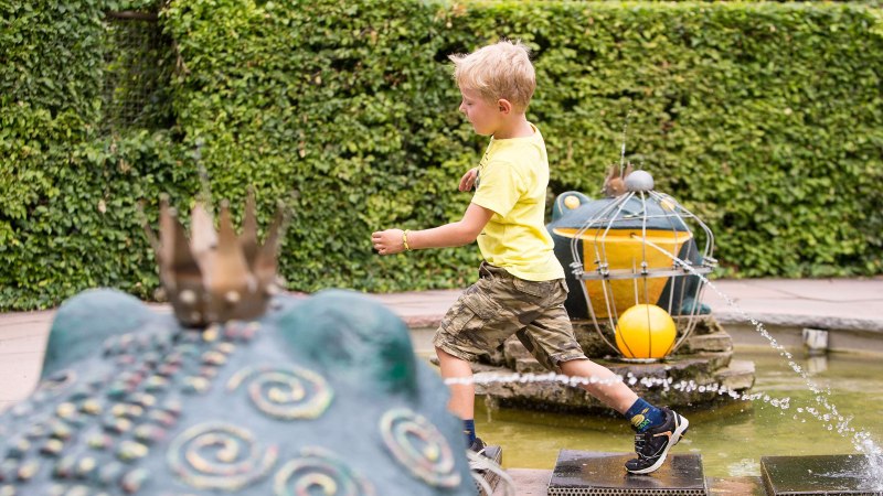Ein Junge in gelbem T-Shirt und Tarnshorts läuft über Steine in einem Märchengarten. Im Hintergrund sind Froschskulpturen und Wasserfontänen zu sehen., © Tourismus & Events Ludwigsburg