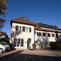 Ein zweistöckiges Hotel mit braunen Fensterläden, umgeben von Bäumen. Ein kleines Auto steht davor. Der Himmel ist klar und blau., © Hotel Bürkle