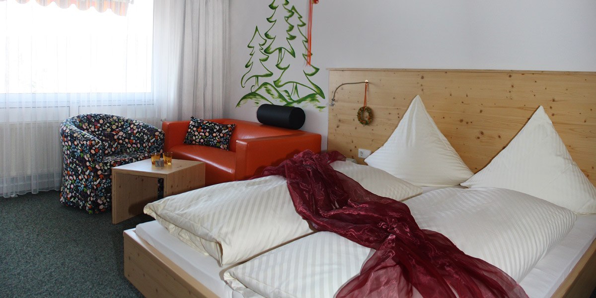 Doppelzimmer mit Holzbett, rotem Sofa, gemustertem Sessel und Wandmalerei von Bäumen. Ein roter Schal liegt auf dem Bett., © Hotel Hochwald