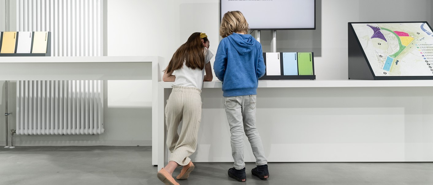 Zwei Kinder betrachten eine Ausstellungstafel in einem modernen Raum mit weißen Wänden und einem Heizkörper., © Stadt Stuttgart / Dominique Brewing
