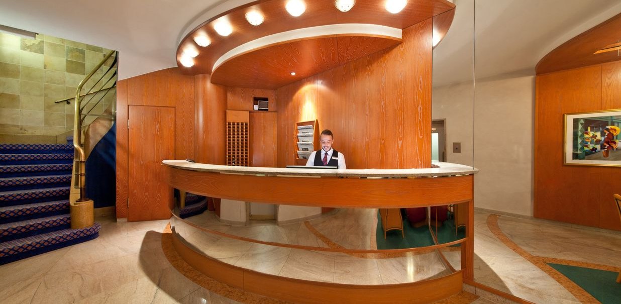 Elegante Hotelrezeption mit Holzverkleidung, beleuchtetem Tresen und einem Mitarbeiter. Links eine Treppe mit blauem Teppich., © Novum Hospitality