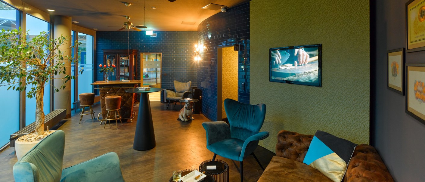 Elegante Zigarrenlounge mit blauen Sesseln, braunem Sofa, Barbereich und stilvoller Dekoration. Ein Fernseher zeigt eine Nahaufnahme von Händen., © Holiday Inn Stuttgart Elegante Zigarrenlounge mit blauen Sesseln, braunem Sofa, Barbereich und stilvoller Dekoration. Ein Fernseher zeigt eine Nahaufnahme von Händen., © Holiday Inn Stuttgart