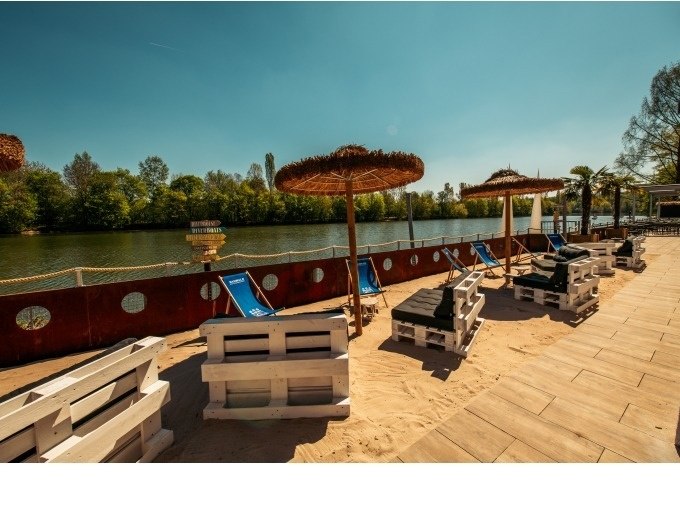 Ein sandiger Strandbereich mit blauen Liegestühlen und Strohdach-Sonnenschirmen am Ufer eines Flusses, umgeben von Bäumen., © Cool-Tours StattReisen Ein sandiger Strandbereich mit blauen Liegestühlen und Strohdach-Sonnenschirmen am Ufer eines Flusses, umgeben von Bäumen., © Cool-Tours StattReisen