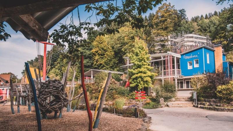 Spielplatz mit buntem Klettergerüst, Holzspänen und einem blauen Gebäude im Hintergrund, umgeben von Bäumen., © Stadt Welzheim Spielplatz mit buntem Klettergerüst, Holzspänen und einem blauen Gebäude im Hintergrund, umgeben von Bäumen., © Stadt Welzheim