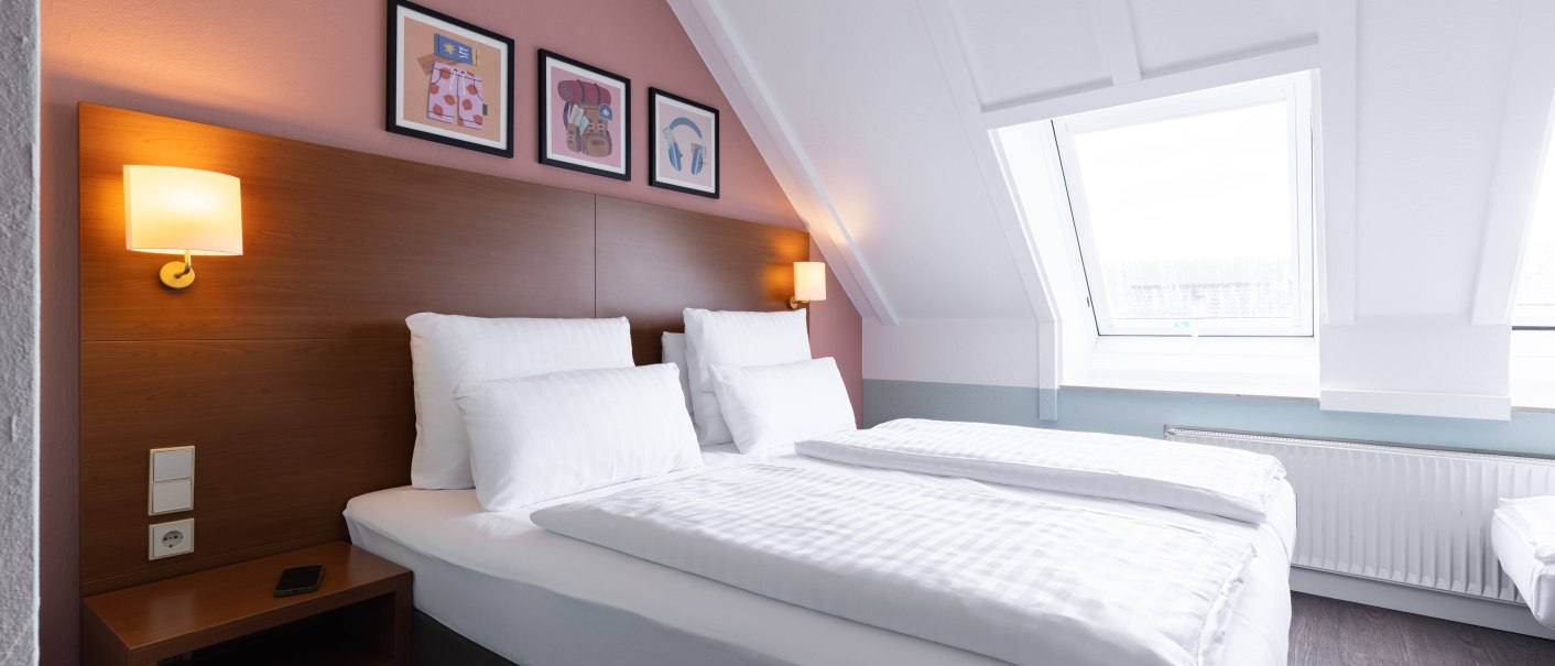 Ein helles Schlafzimmer mit Doppelbett, wei&szlig;en Bettlaken und Holzkopfteil. An der Wand h&auml;ngen drei Bilder, daneben sind Wandlampen angebracht., &copy; Novum  Hospitality