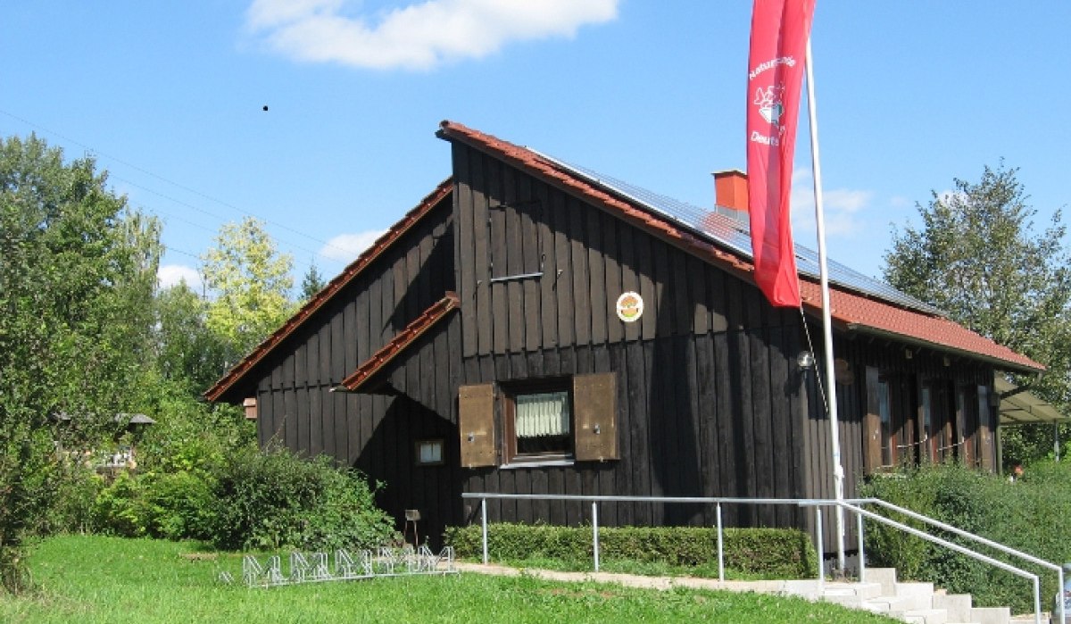 Holzhaus mit rotem Dach und Solarpanelen, rote Fahne daneben. Umgeben von grüner Wiese und Bäumen, blauer Himmel im Hintergrund., © Naturfreunde Holzgerlingen/Altdorf