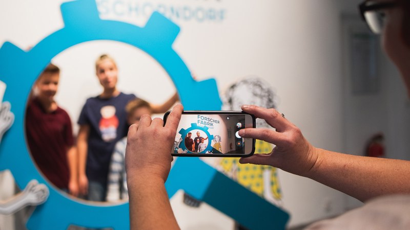 Eine Person fotografiert drei Kinder durch einen blauen, zahnradförmigen Rahmen mit einem Smartphone. Im Hintergrund sind Zeichnungen zu sehen., © Danijel Grbic
