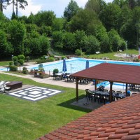 Ein Freibad mit Schwimmbecken, umgeben von Bäumen. Es gibt Sitzbereiche mit Sonnenschirmen und einen überdachten Pavillon., © FVFS