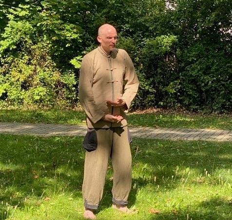 Ein Mann in traditioneller Qigong-Kleidung steht barfuß auf einer Wiese und führt eine Übung durch. Im Hintergrund sind Bäume und Büsche zu sehen., © Daniel Steinbauer Ein Mann in traditioneller Qigong-Kleidung steht barfuß auf einer Wiese und führt eine Übung durch. Im Hintergrund sind Bäume und Büsche zu sehen., © Daniel Steinbauer