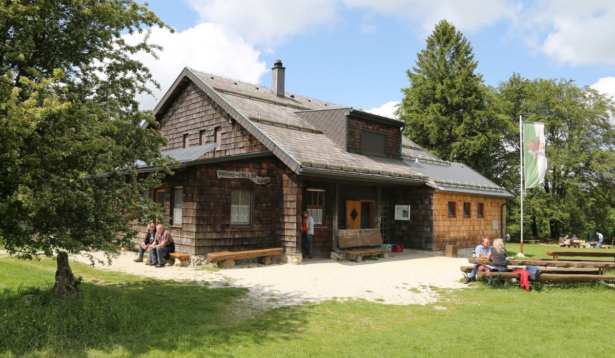 Das Franz Keller Haus ist ein rustikales Holzhaus in einer grünen Umgebung. Menschen sitzen auf Bänken, während eine Fahne im Wind weht., © Fotograf Walter Laible Das Franz Keller Haus ist ein rustikales Holzhaus in einer grünen Umgebung. Menschen sitzen auf Bänken, während eine Fahne im Wind weht., © Fotograf Walter Laible