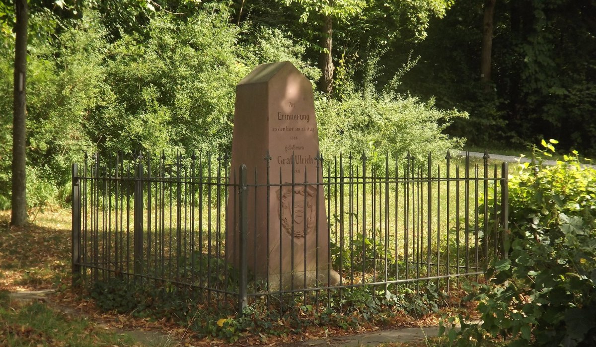 Ein Gedenkstein für Graf Ulrich, umgeben von einem Metallzaun, steht in einem bewaldeten Gebiet. Der Stein trägt eine Inschrift zur Erinnerung., © Natur.Nah. Schönbuch & Heckengäu Ein Gedenkstein für Graf Ulrich, umgeben von einem Metallzaun, steht in einem bewaldeten Gebiet. Der Stein trägt eine Inschrift zur Erinnerung., © Natur.Nah. Schönbuch & Heckengäu