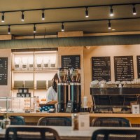 Modernes Caf&eacute; mit Theke, Kaffeemaschine und Speisekarten an der Wand. Pflanzen im Vordergrund, gem&uuml;tliche Atmosph&auml;re., &copy; Maison Karl, Stuttgart