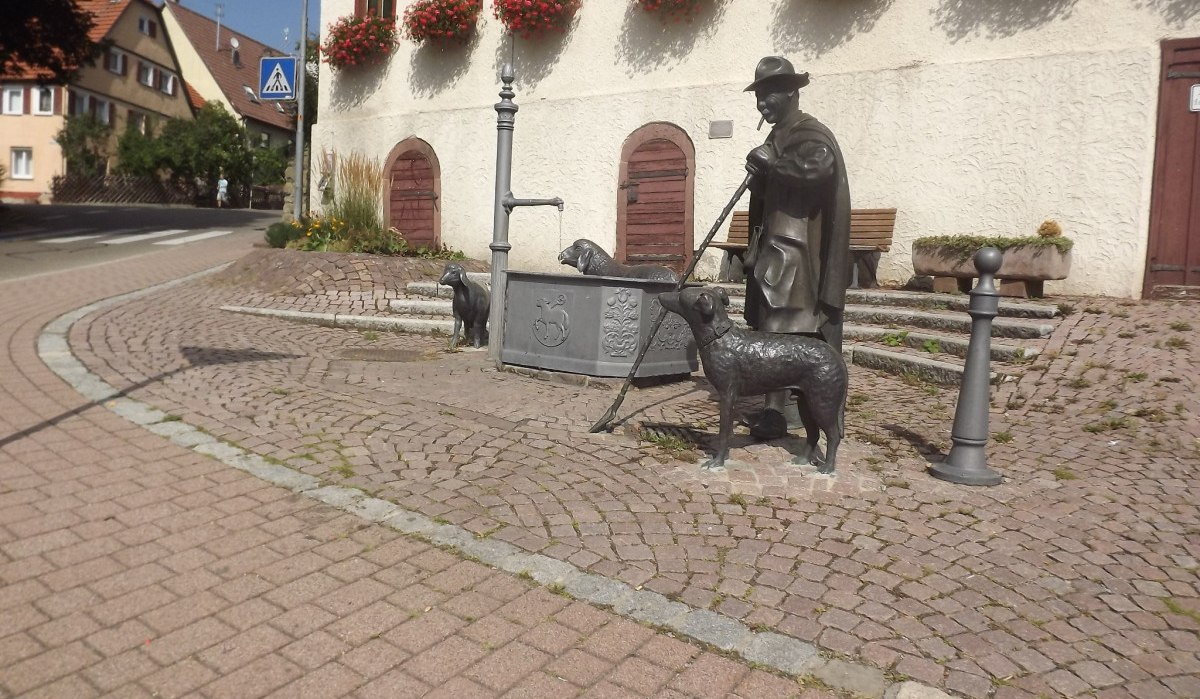 Bronzestatue eines Mannes mit Schafen an einem Brunnen auf einer gepflasterten Straße. Im Hintergrund ein Gebäude mit roten Blumen an den Fenstern., © Natur.Nah. Schönbuch & Heckengäu Bronzestatue eines Mannes mit Schafen an einem Brunnen auf einer gepflasterten Straße. Im Hintergrund ein Gebäude mit roten Blumen an den Fenstern., © Natur.Nah. Schönbuch & Heckengäu