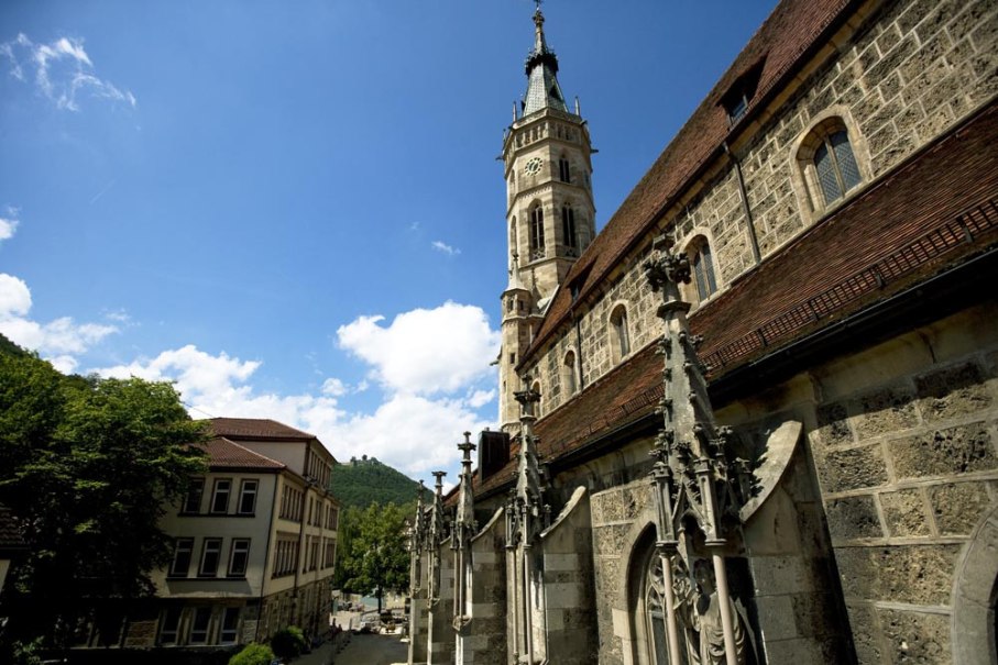 Eine Kirche mit einem hohen Turm und gotischen Elementen, umgeben von Bäumen und Gebäuden, unter einem klaren blauen Himmel., © Bad Urach Tourismus