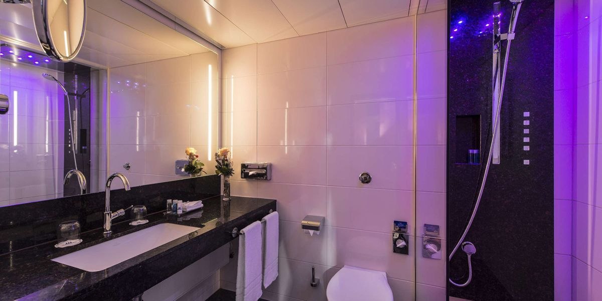 Modernes Badezimmer mit dunklem Waschtisch, großem Spiegel, Dusche und lila Beleuchtung. Handtücher und Blumen dekorieren den Raum., © Maritim Hotelgesellschaft mbH