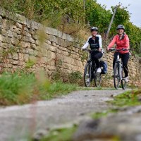 Zwei Frauen radeln lächelnd auf einem Weg neben einer alten Steinmauer, umgeben von grüner Vegetation., © Kraichgau Stromberg Tourismus e.V.-Christian Ernst Zwei Frauen radeln lächelnd auf einem Weg neben einer alten Steinmauer, umgeben von grüner Vegetation., © Kraichgau Stromberg Tourismus e.V.-Christian Ernst